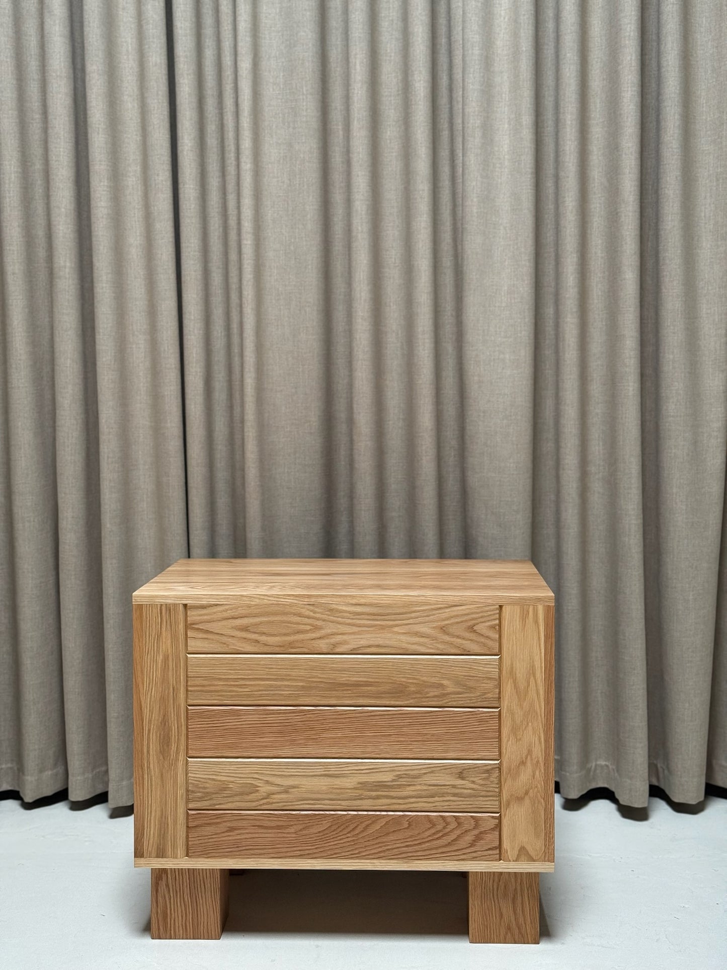 Wooden nightstand with three drawers against a gray curtain background