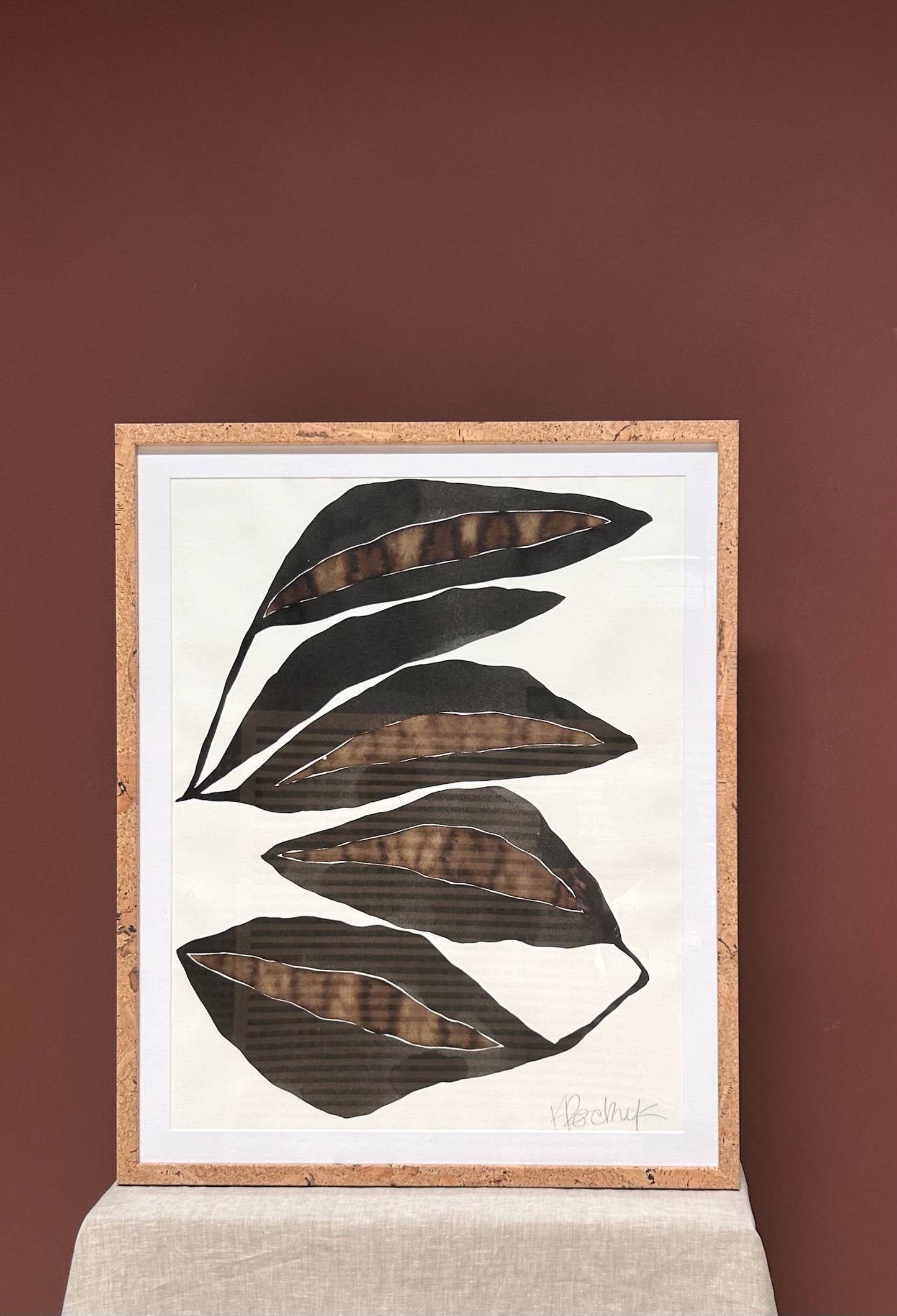 Framed artwork with black leaf shapes on a white background against a brown wall.